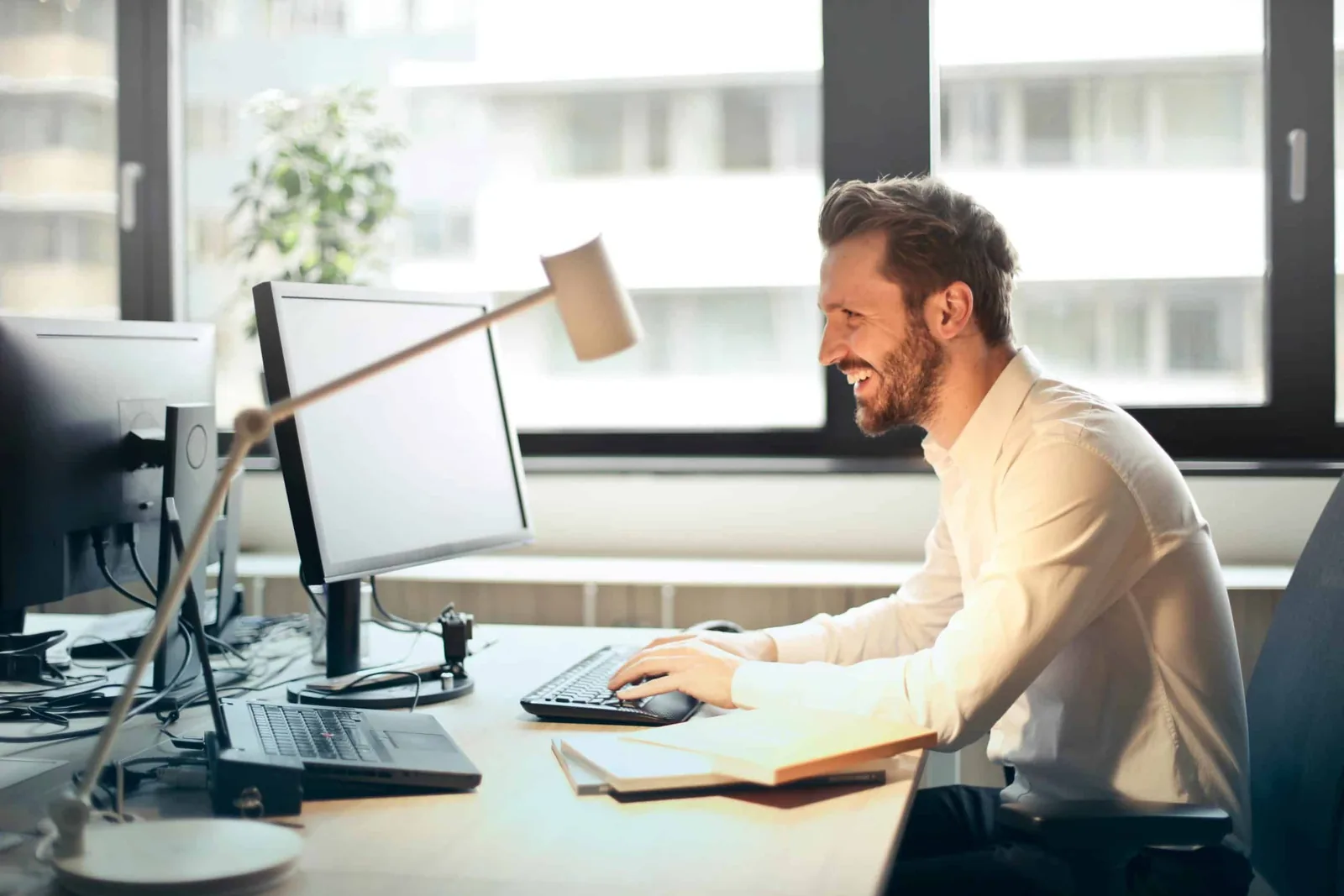 Man achter twee schermen aan een bureau bij het raam, met papieren naast zich